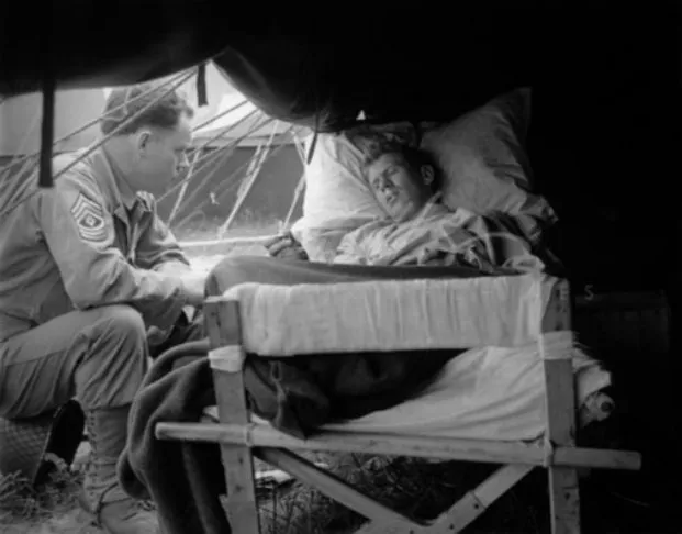 Black-and-white photograph inside a hospital tent: a soldier in uniform sits beside a camp bed and leans towards a patient who lies propped on pillows with his eyes closed.