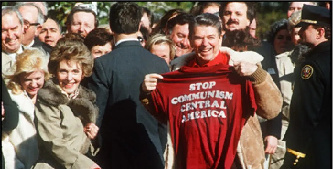 Photograph of Ronald Reagan in a crowd, holding up a red T-shirt printed “Stop Communism Central America”. Nancy Reagan stands beside him, smiling.