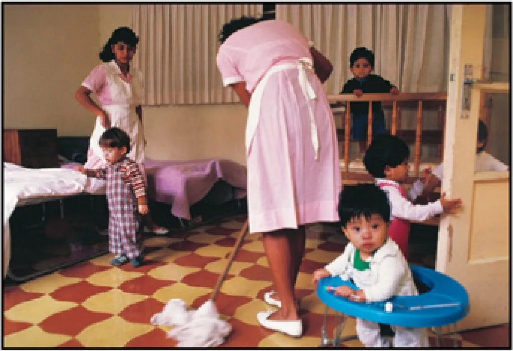 Photograph of a room with a patterned tile floor where women in pink uniforms look after several toddlers. One woman mops the floor; a child sits in a blue baby walker in the foreground.