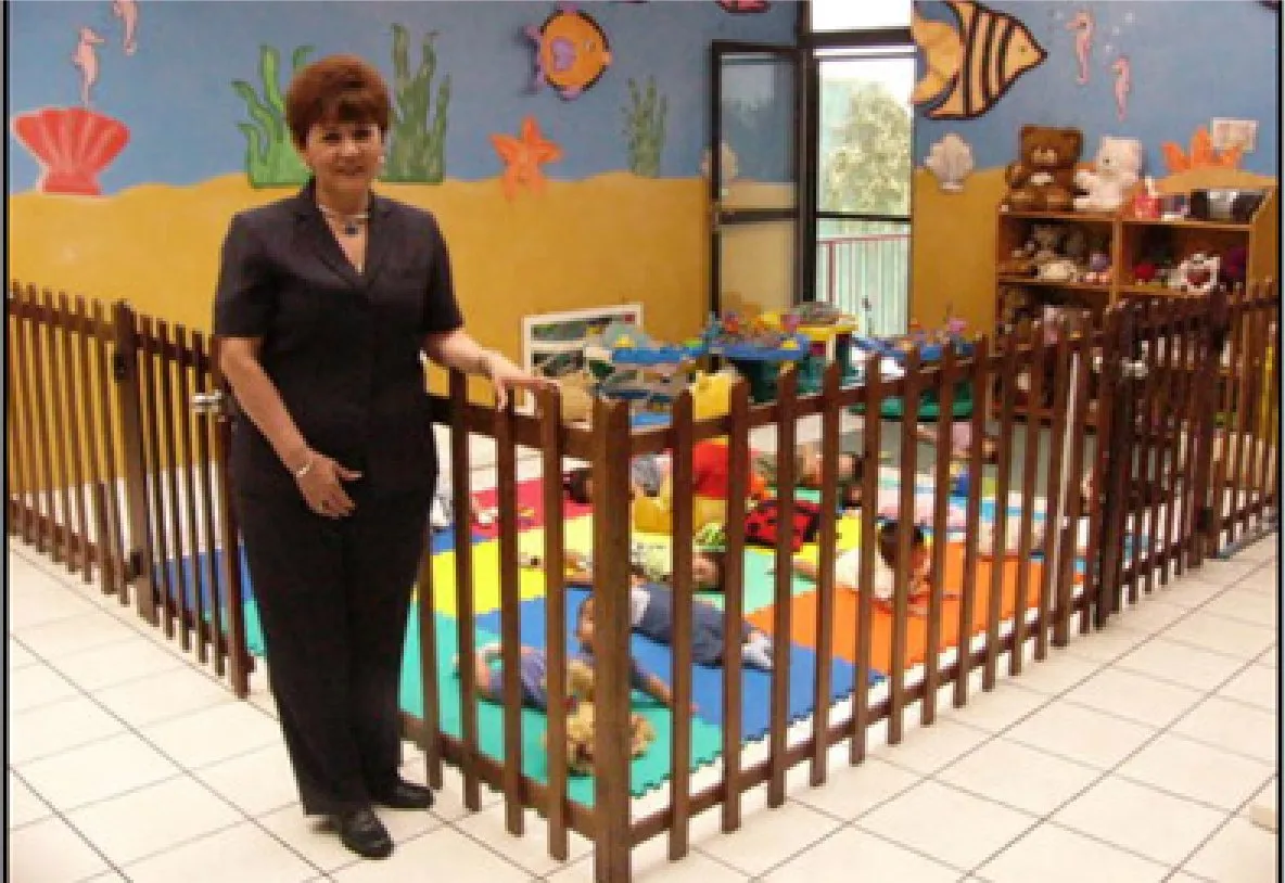 Photograph of a woman in a dark suit standing beside a wooden playpen fence in a nursery painted with sea creatures. Babies lie on coloured mats inside the fence.