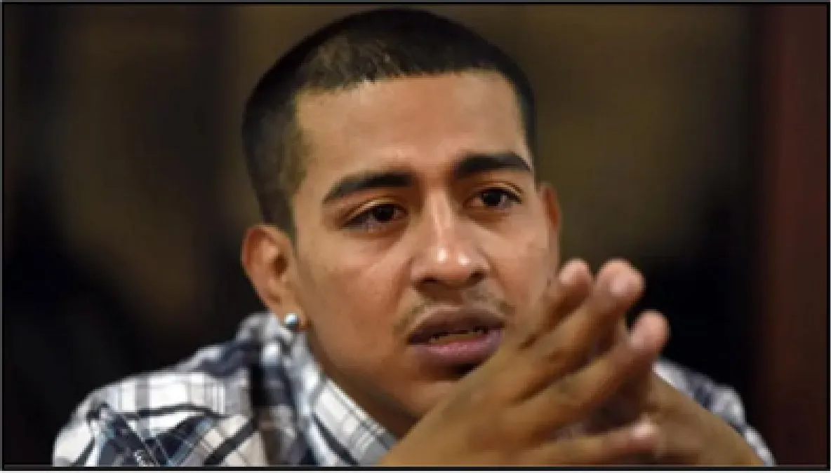 Close-up photograph of a young man in a plaid shirt with his hands pressed together in front of him, looking to one side with a serious expression.