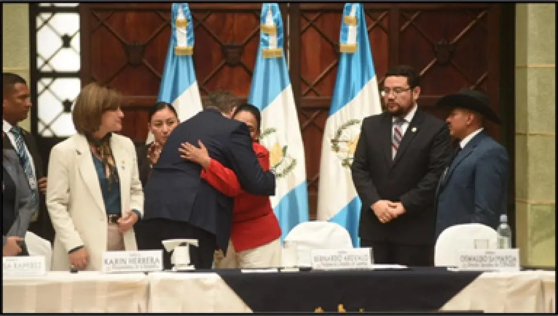 Photograph of a formal ceremony in front of Guatemalan flags. A man in a dark suit embraces a woman dressed in red while officials standing on either side look on.